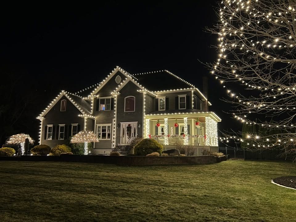 Two-story home with warm white C9 roofline, lit porch columns, garland and wrapped bushes — professional Christmas light installation